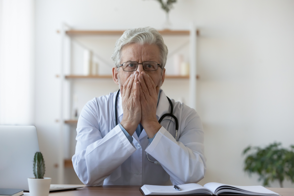 Confused old mature doctor therapist in glasses and white coat covering mouth with hands